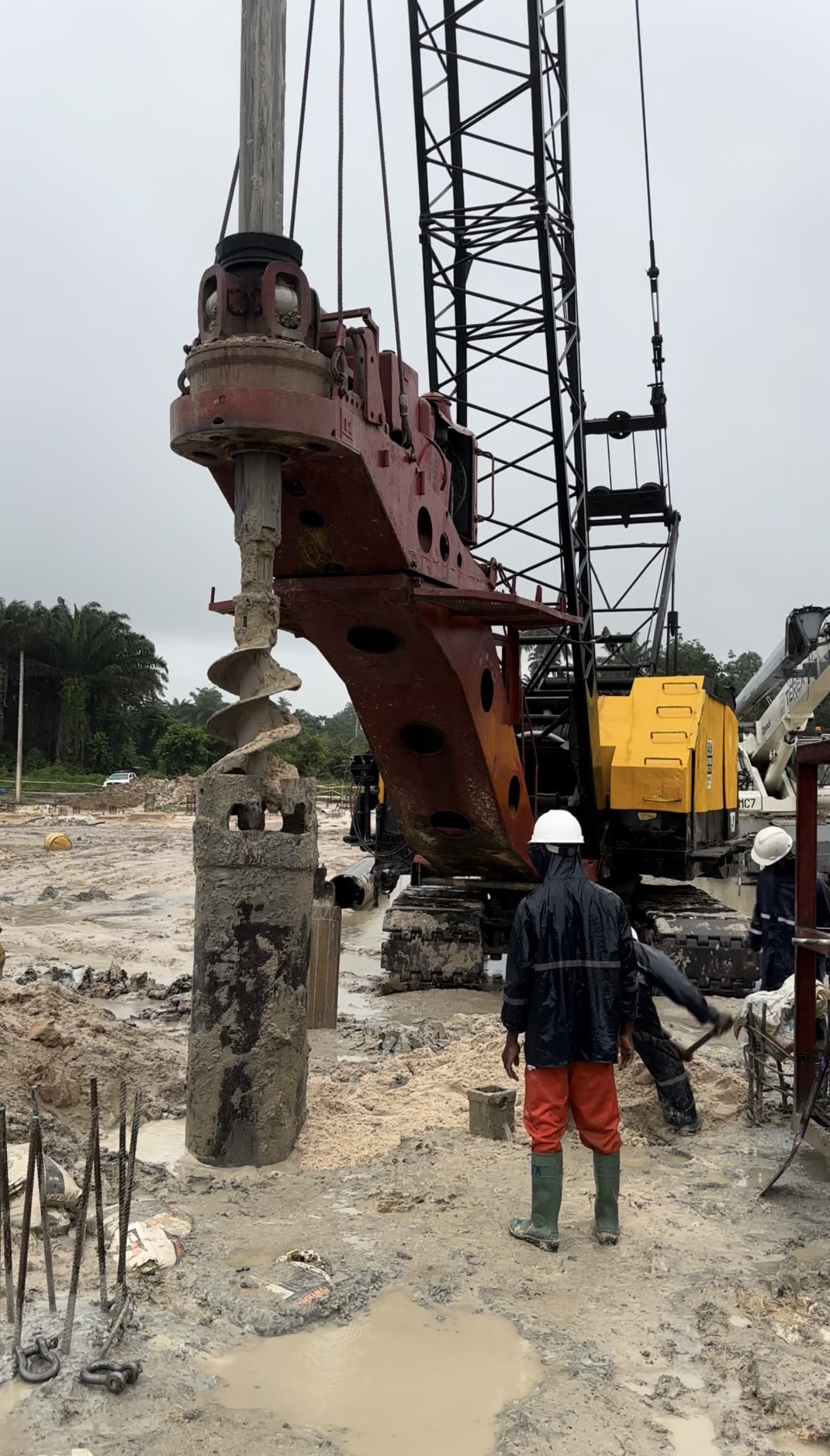 A rotary rig lifting a soil-filled auger clear of the bore beside a set steel casing, crew working at ground level