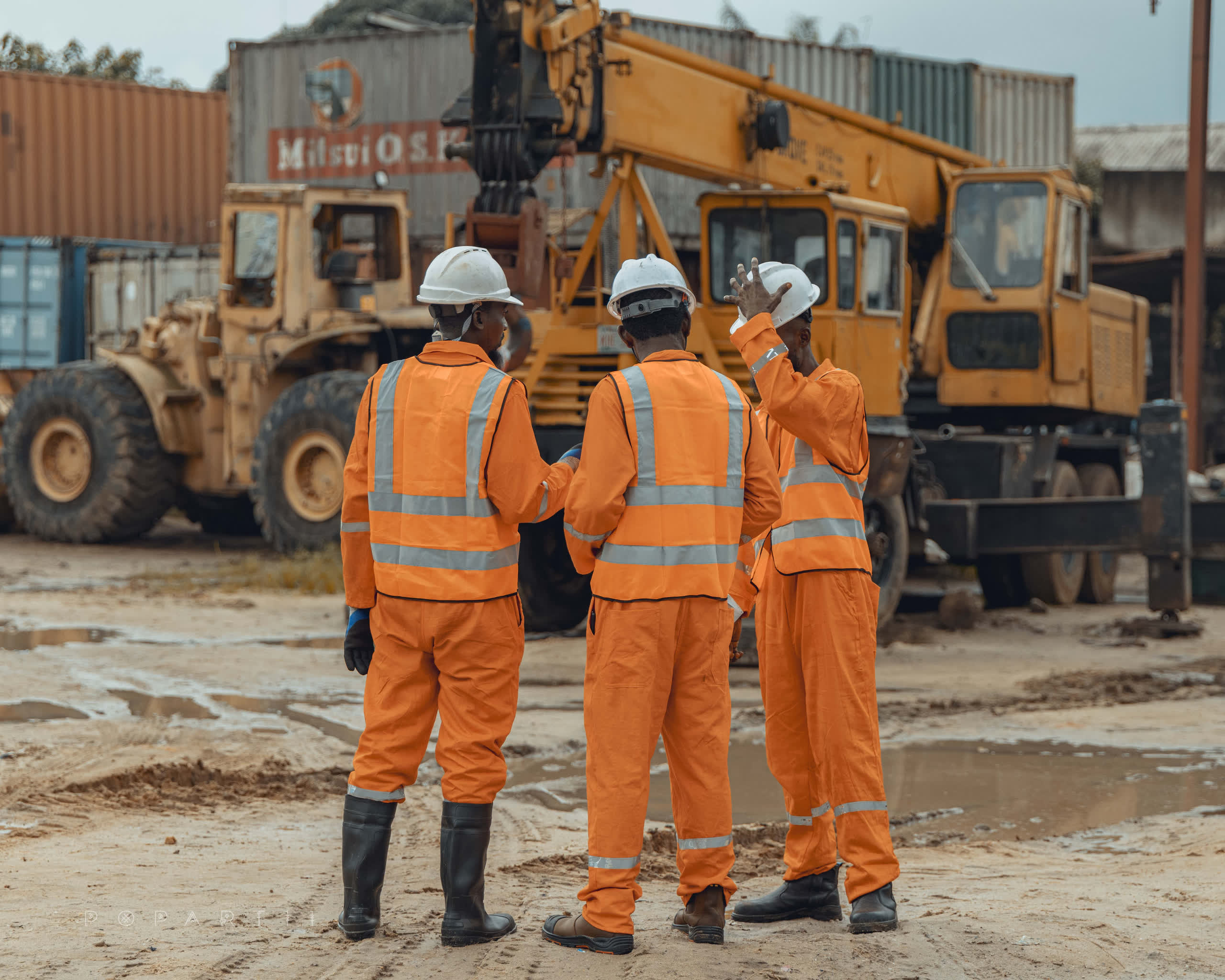 Three crew members in orange coveralls conferring in front of shipping containers, a yellow mobile crane and wheel loader behind
