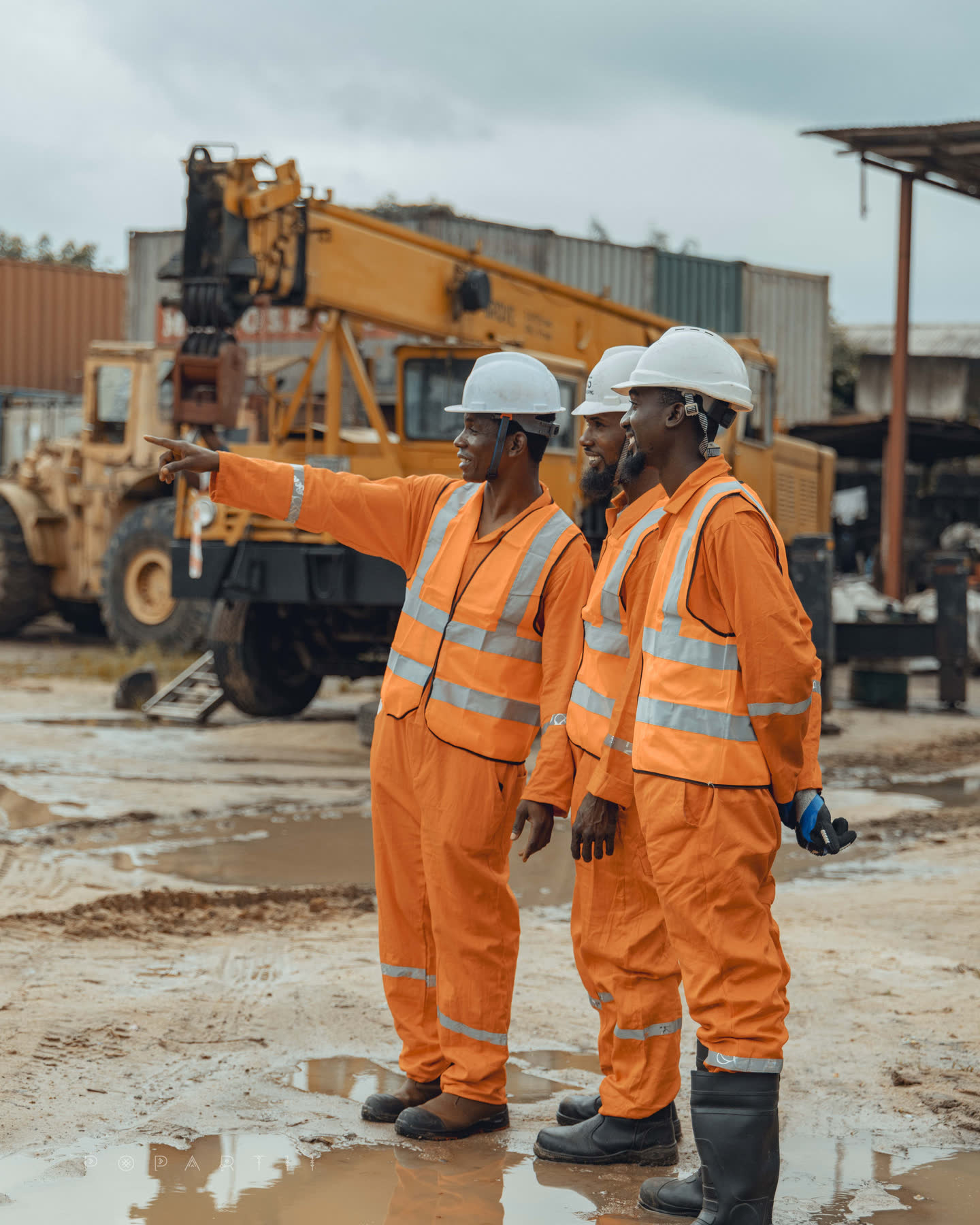 A crew member in orange coveralls pointing across the yard while two colleagues look on, a wheel loader and crane behind them