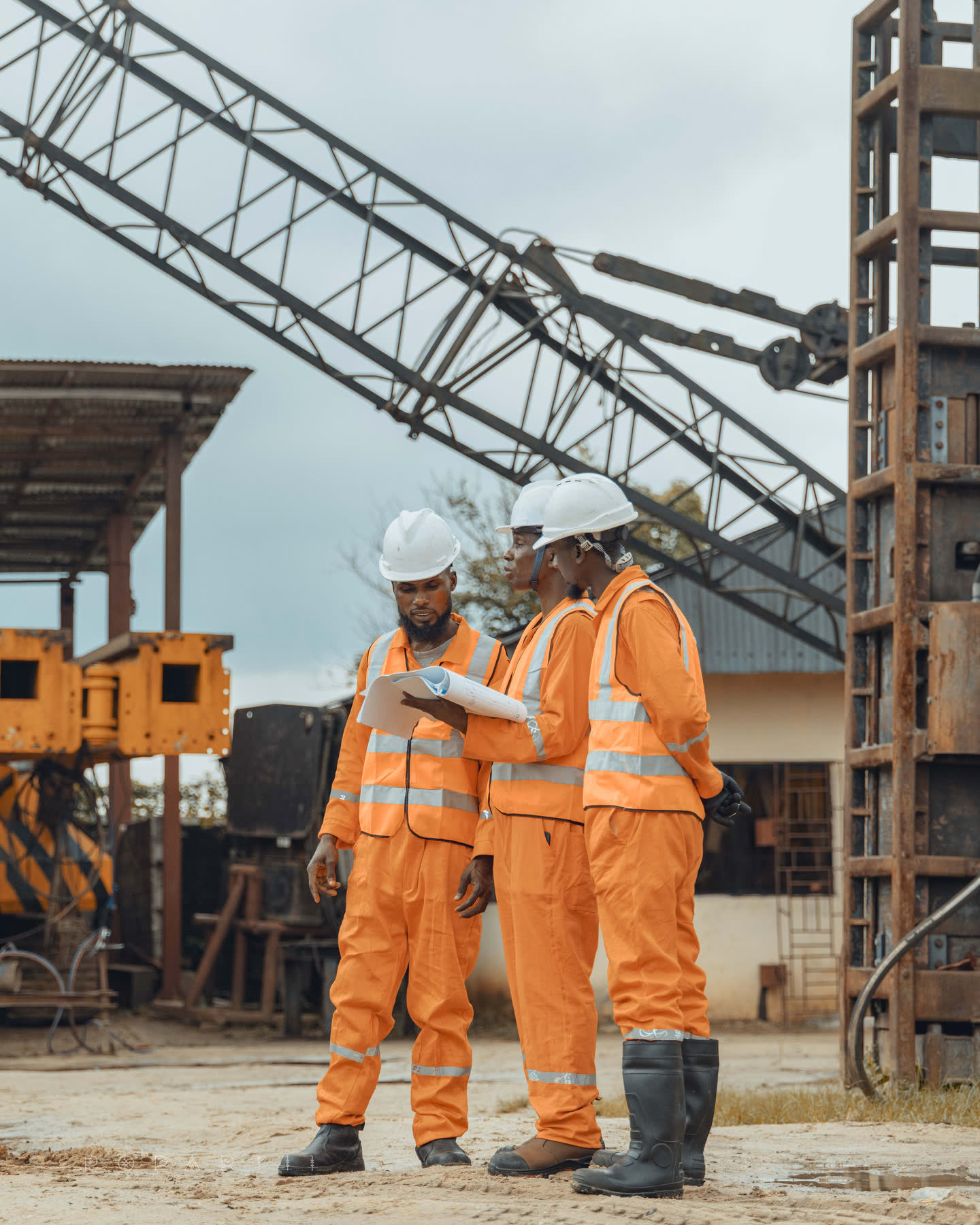 Three crew members in orange coveralls reading a drawing together beneath a lattice crane boom, piling leads standing behind them
