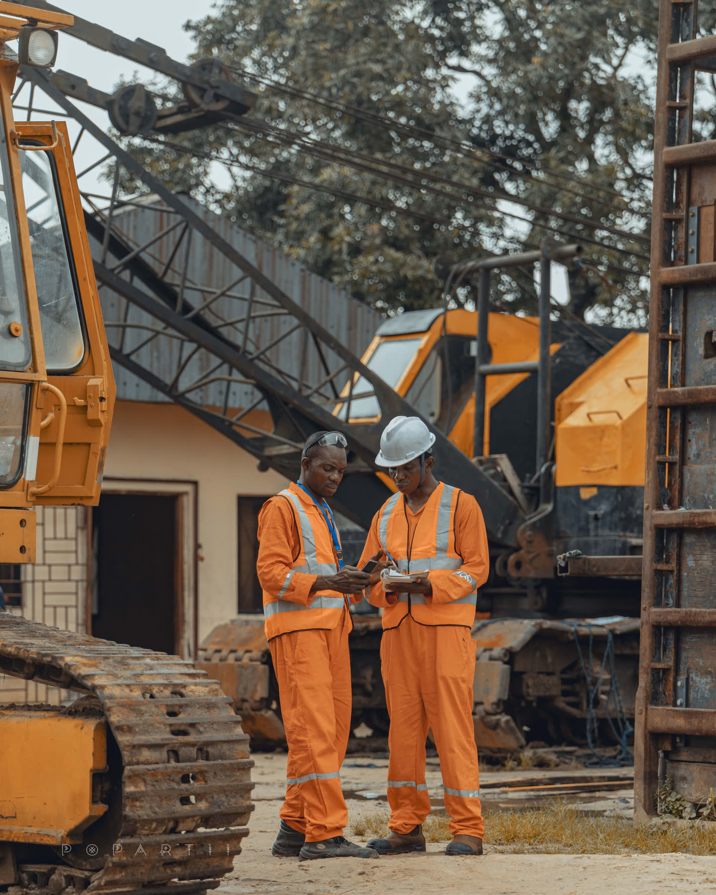 Two crew members in orange coveralls and hard hats reading notes together beside the tracks of a crawler crane in the yard