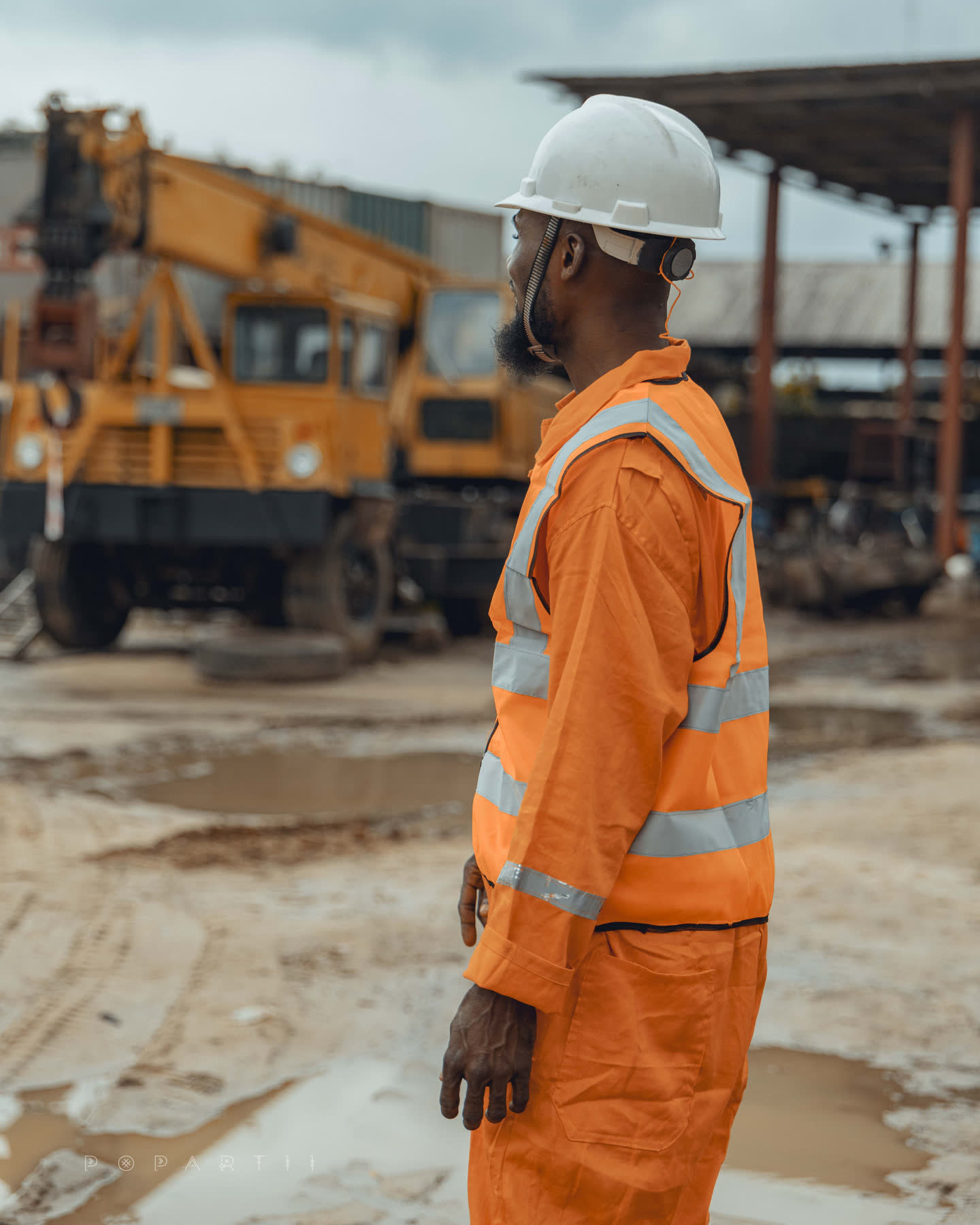 A crew member in orange high-visibility coveralls and a white hard hat seen in profile, wheeled cranes standing on wet ground behind