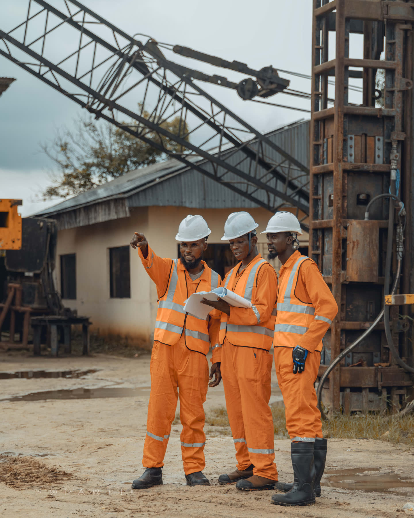 Three crew members in orange coveralls and white hard hats standing beneath a crane boom and drop-hammer leads, one pointing off frame