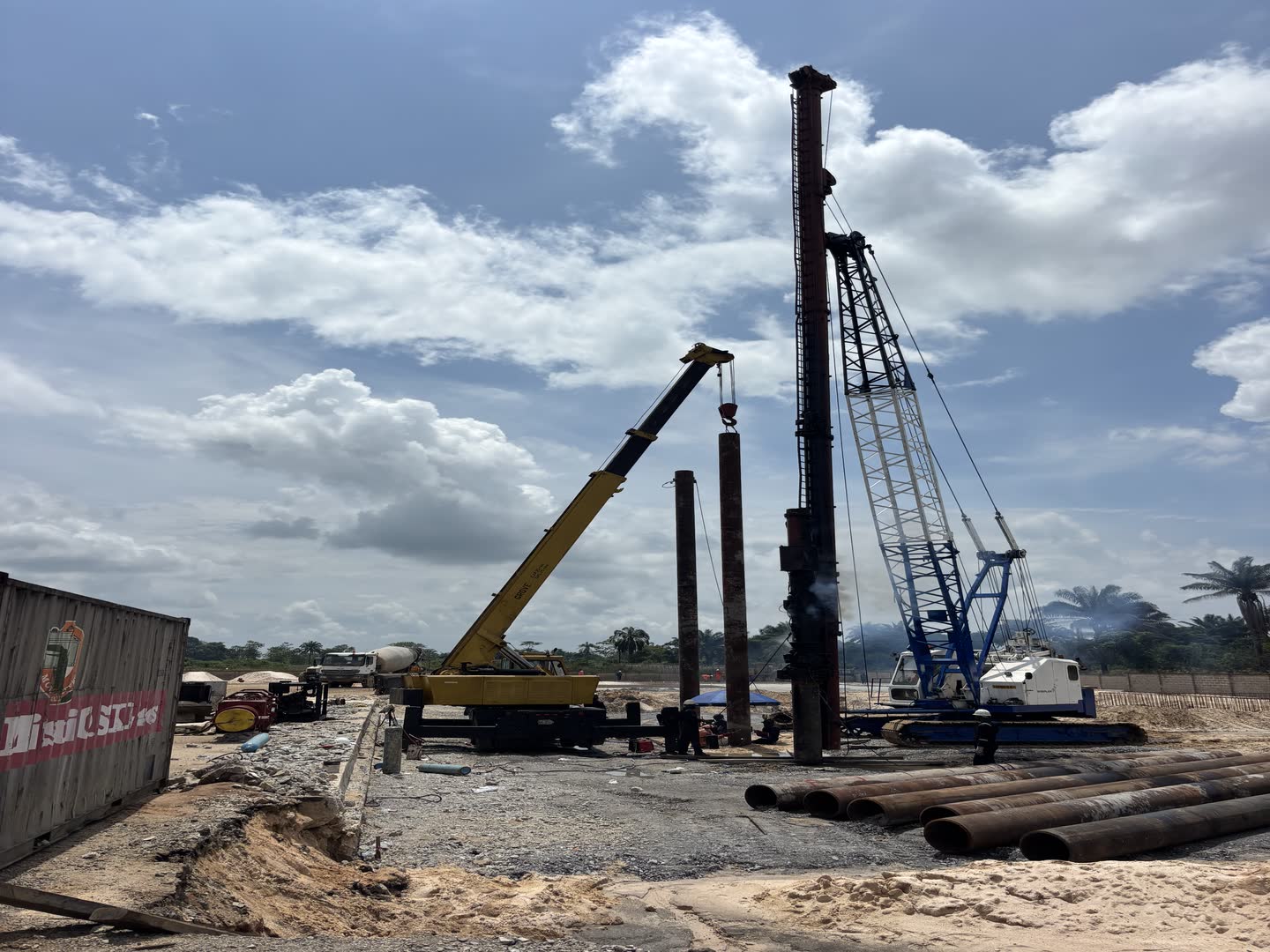 A crawler crane with piling leads raised beside pitched steel casings, a service crane and truck alongside on a gravel platform