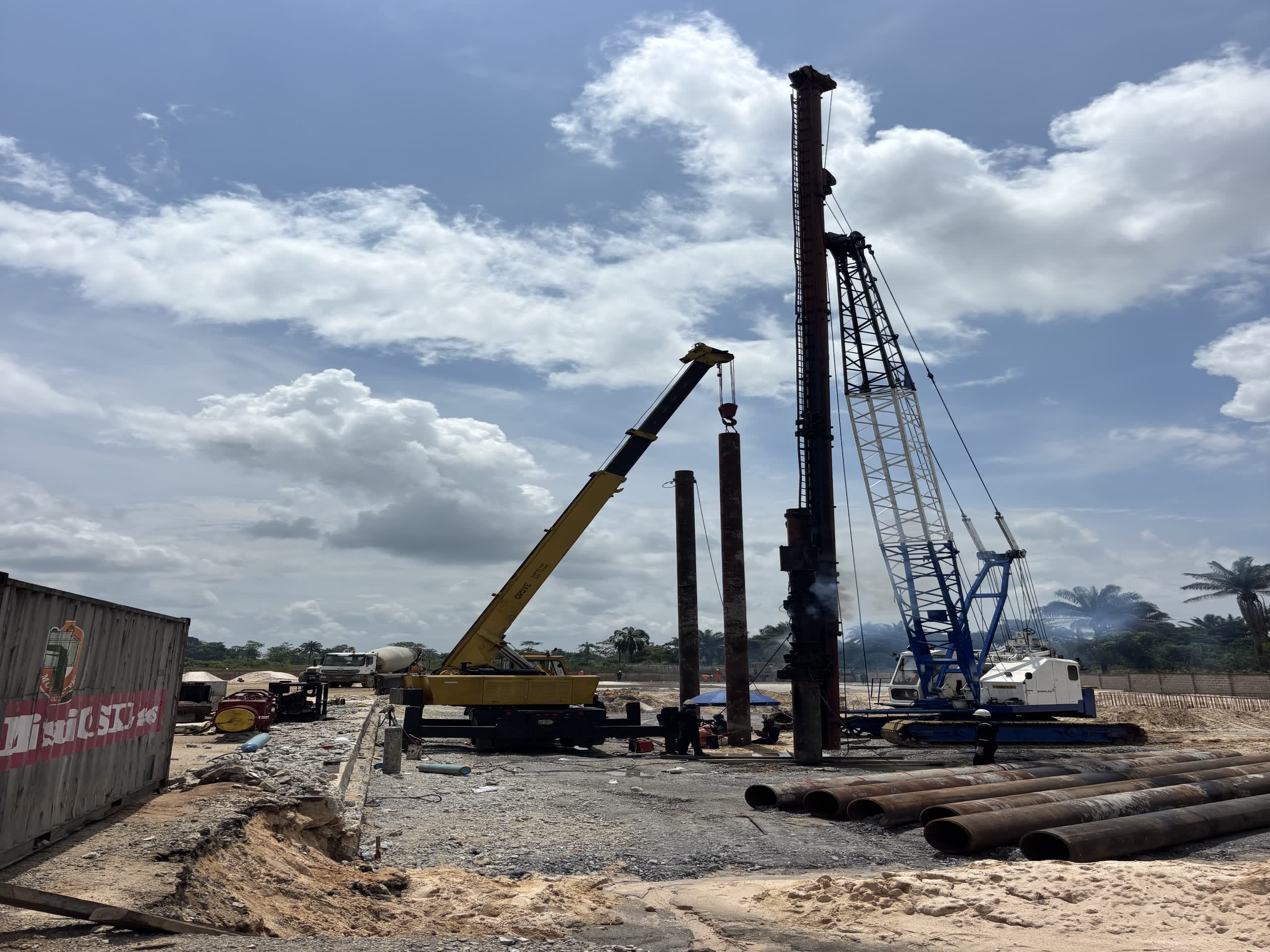 A crawler crane with piling leads raised beside pitched steel casings, a service crane and truck alongside on a gravel platform