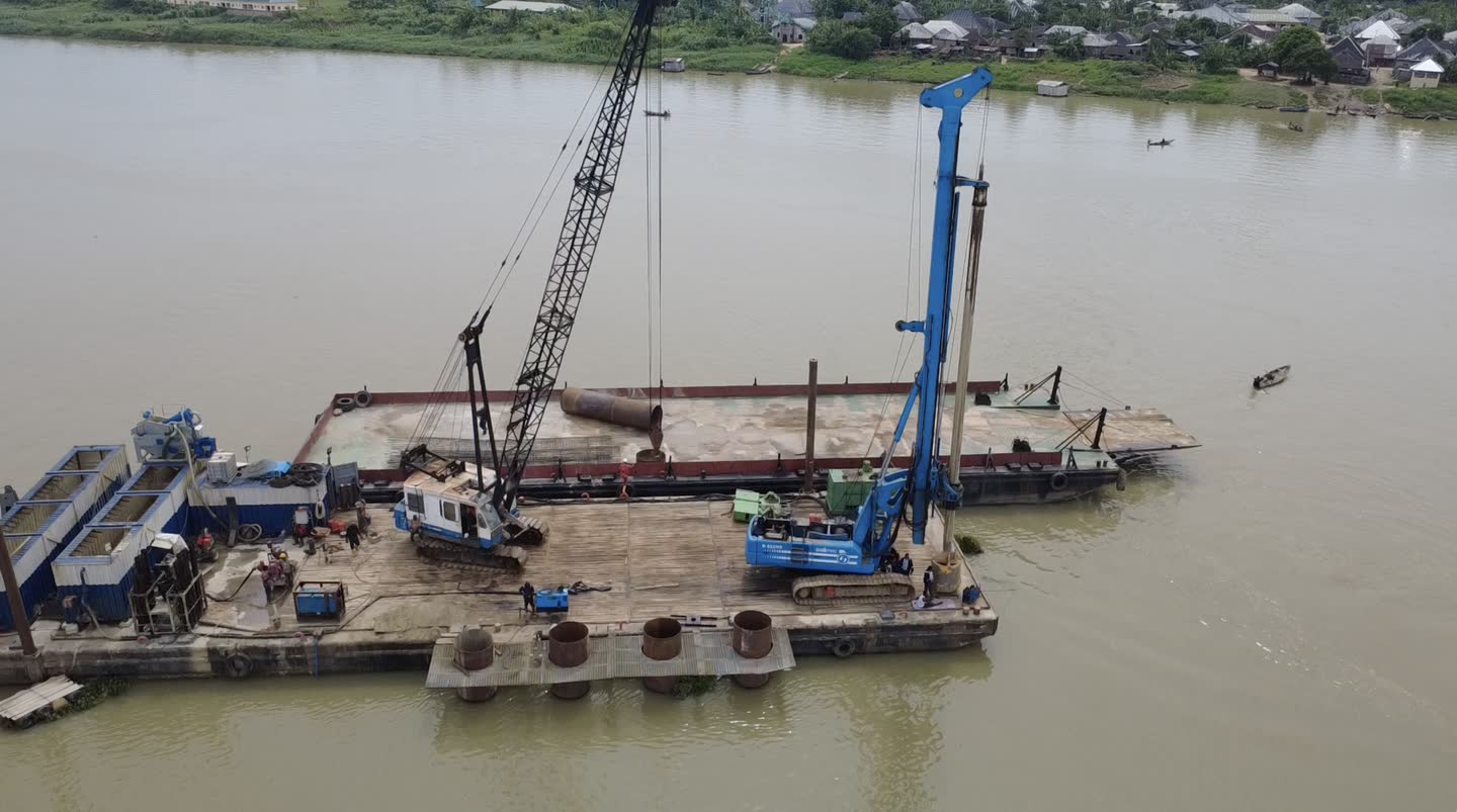 Aerial view of a lattice-boom crane and a blue tracked piling rig working from a moored barge on a wide river, a village on the far bank