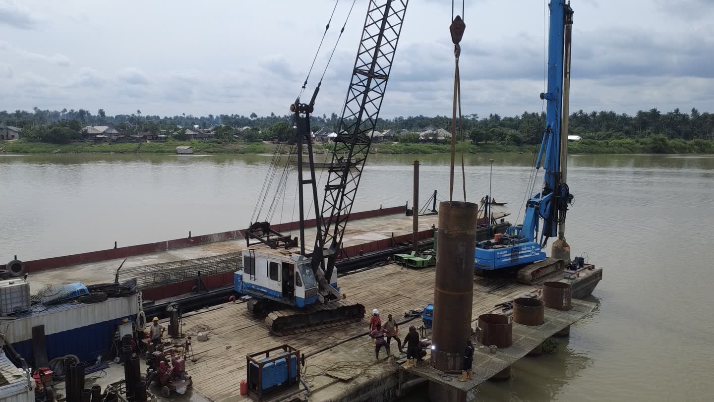 A steel pile casing suspended from a crane hook over a barge deck while crew in hard hats steady it, a blue piling rig alongside