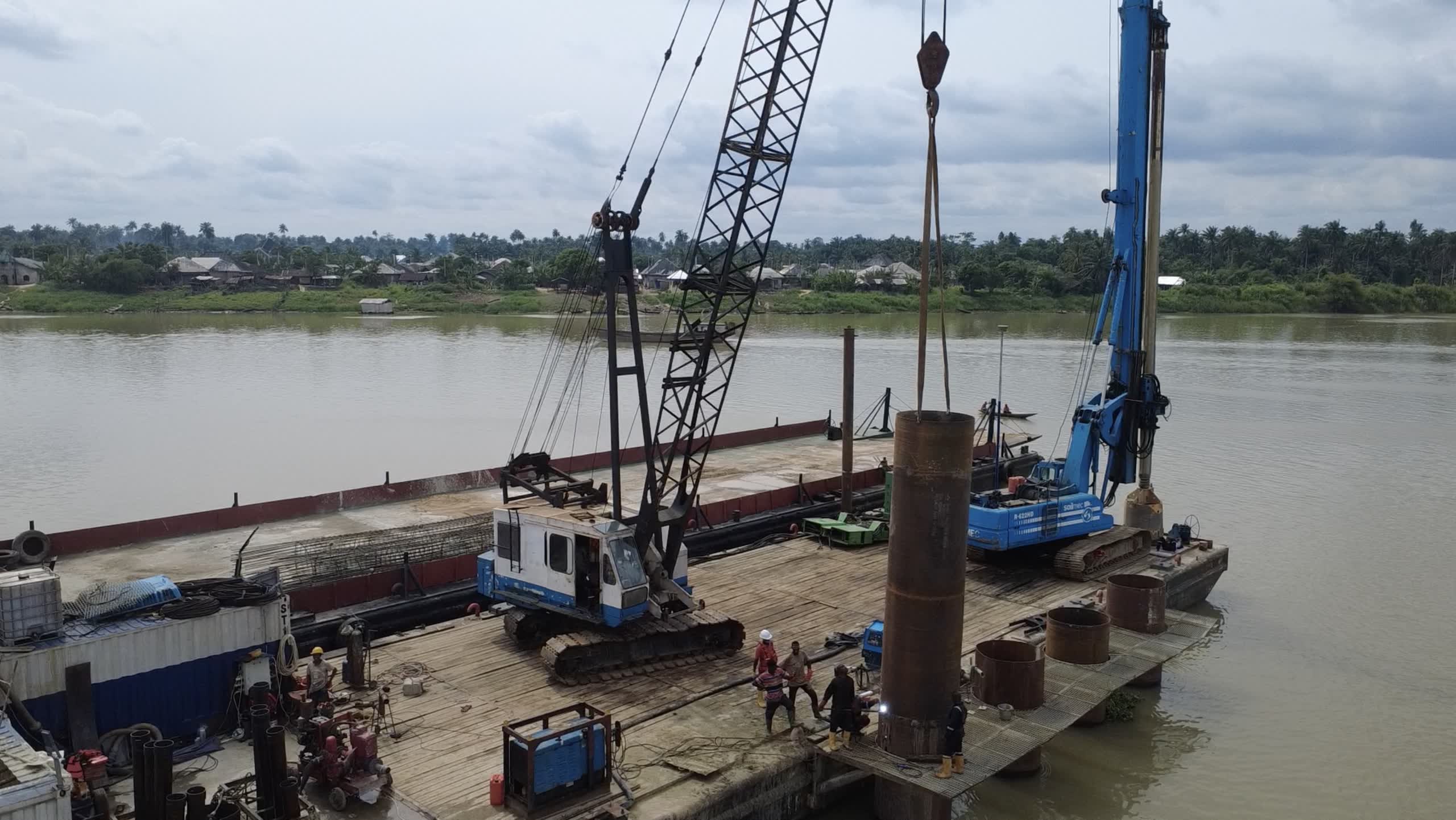 A steel pile casing suspended from a crane hook over a barge deck while crew in hard hats steady it, a blue piling rig alongside