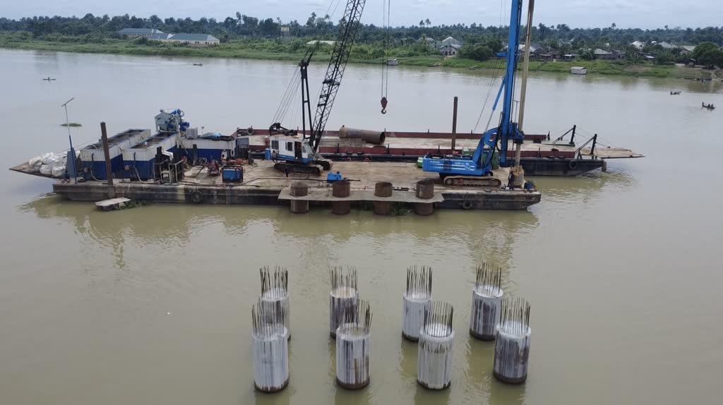 Eight completed cased piles standing out of a river with reinforcement bars projecting from each head, the piling barge moored behind them