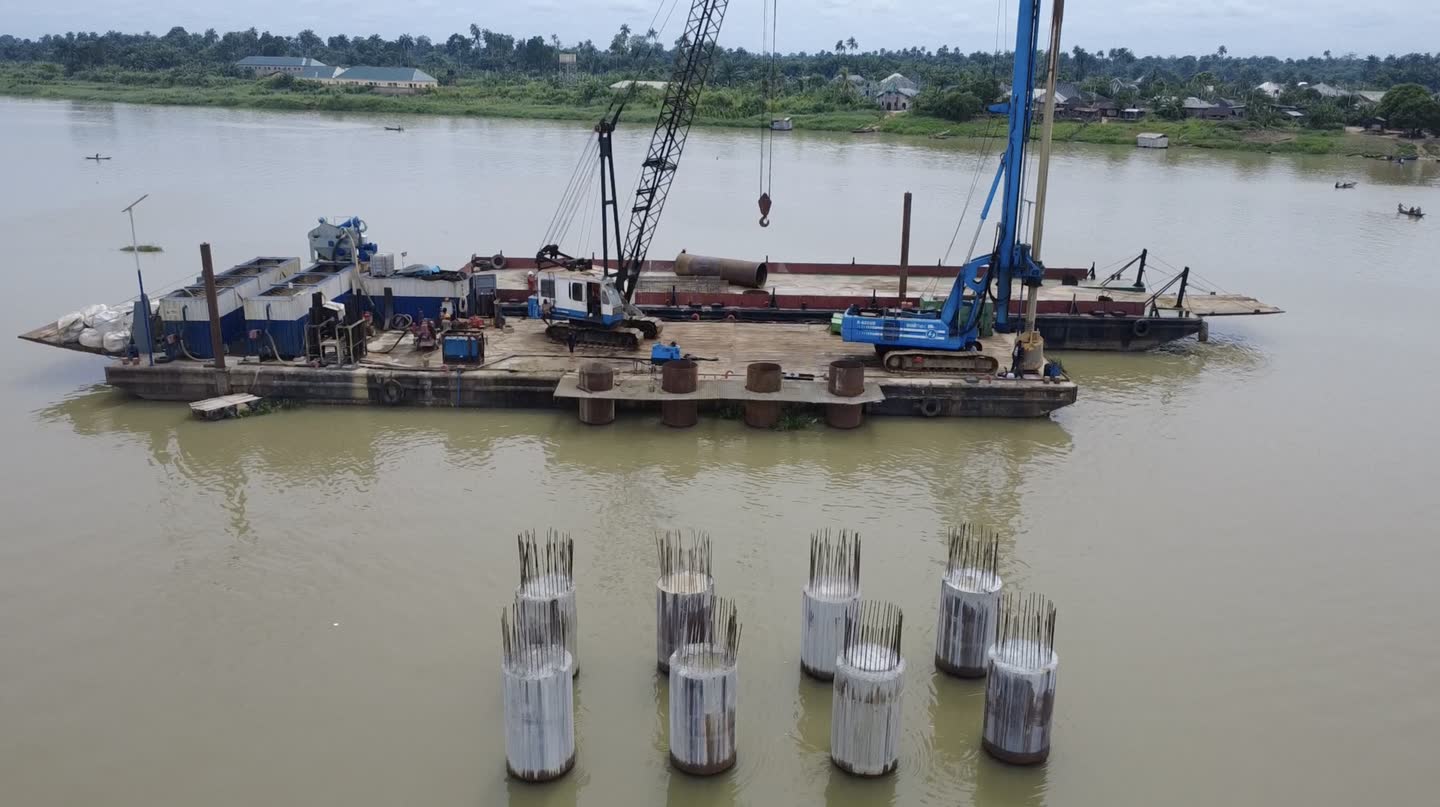 Eight completed cased piles standing out of a river with reinforcement bars projecting from each head, the piling barge moored behind them