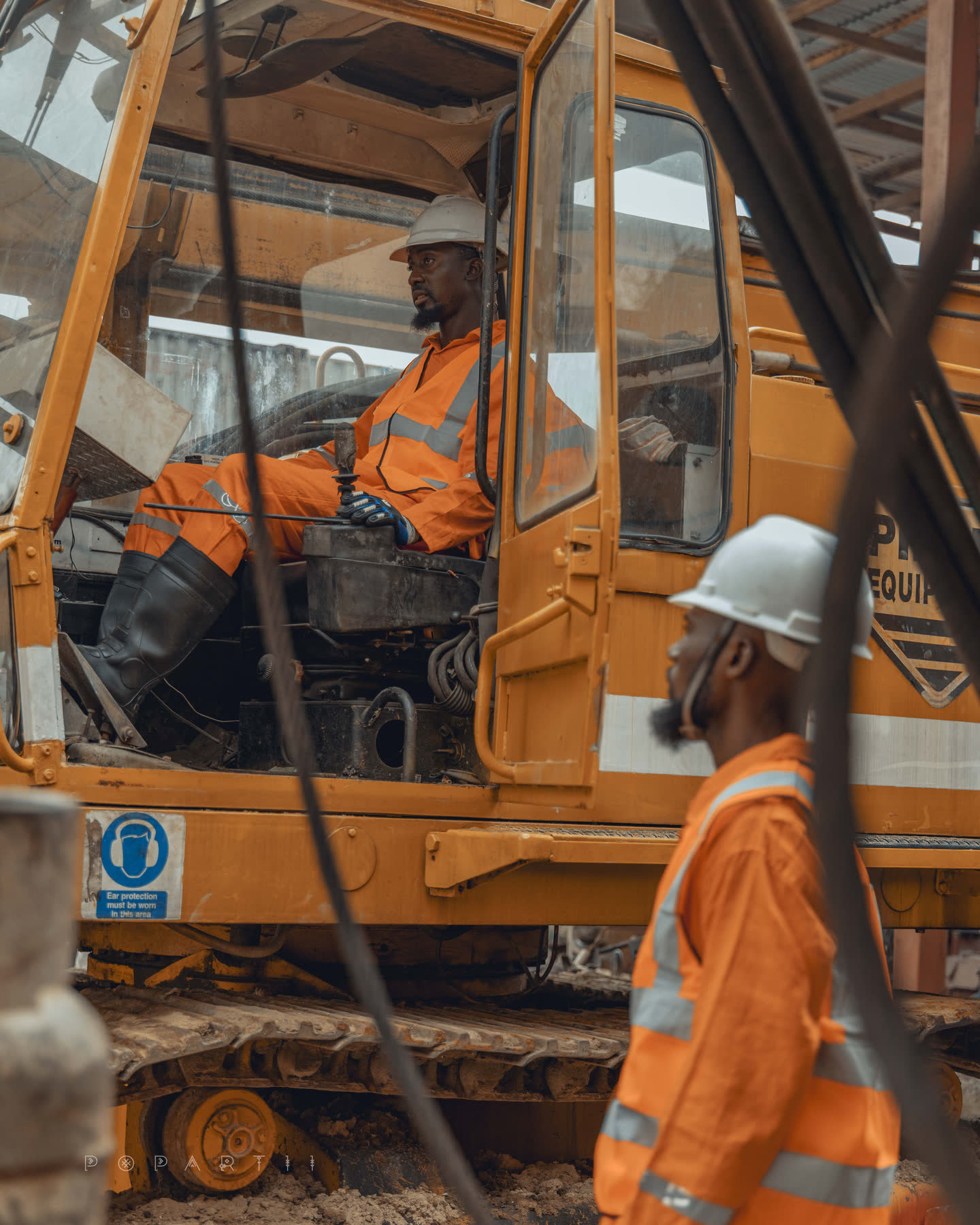 An operator in orange coveralls and a white hard hat seated at the controls of a crawler crane, a second crew member passing in the foreground