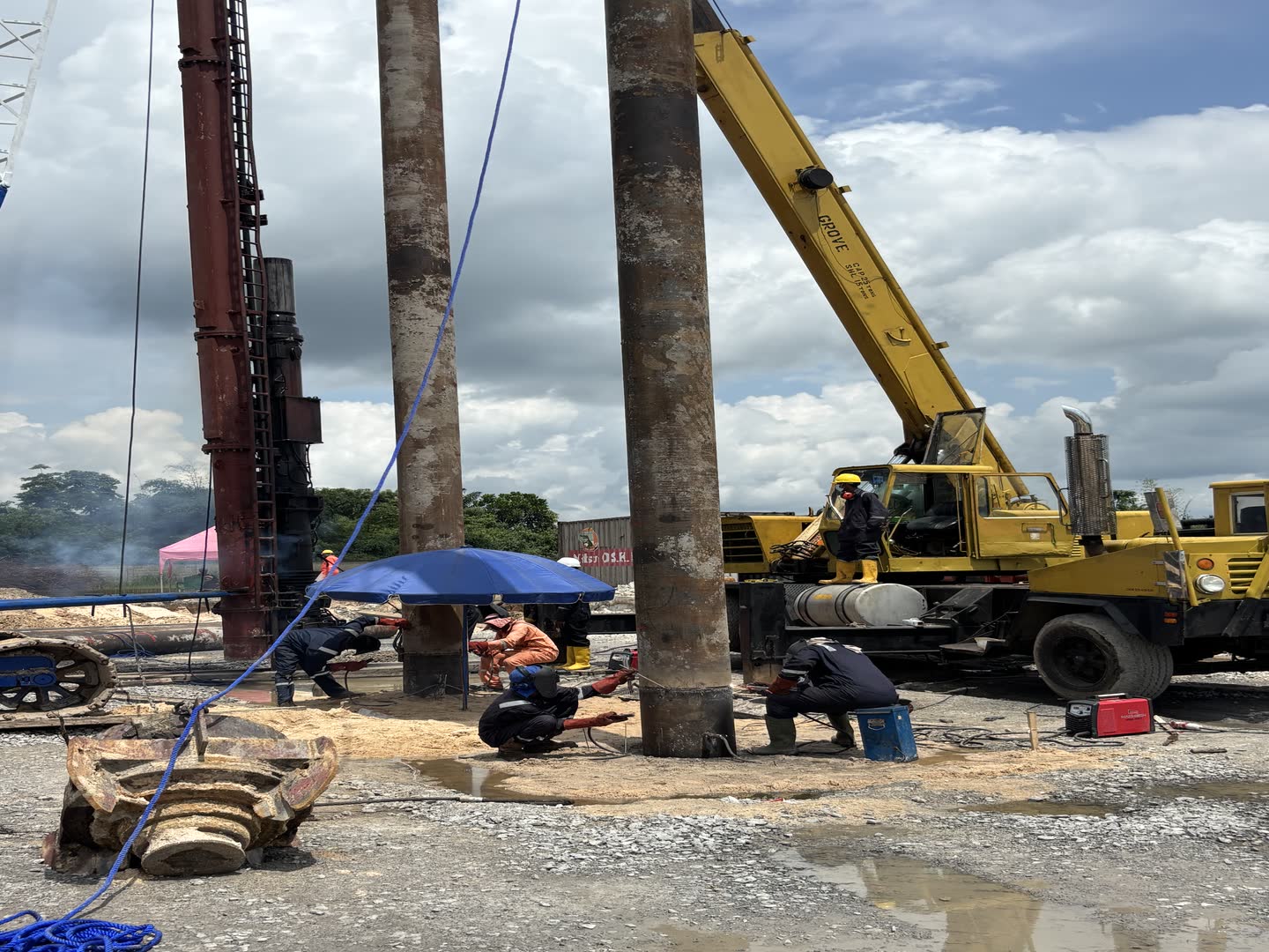 A tracked piling rig and a yellow service crane working beneath tall cumulus cloud, steel pile sections laid out on the ground