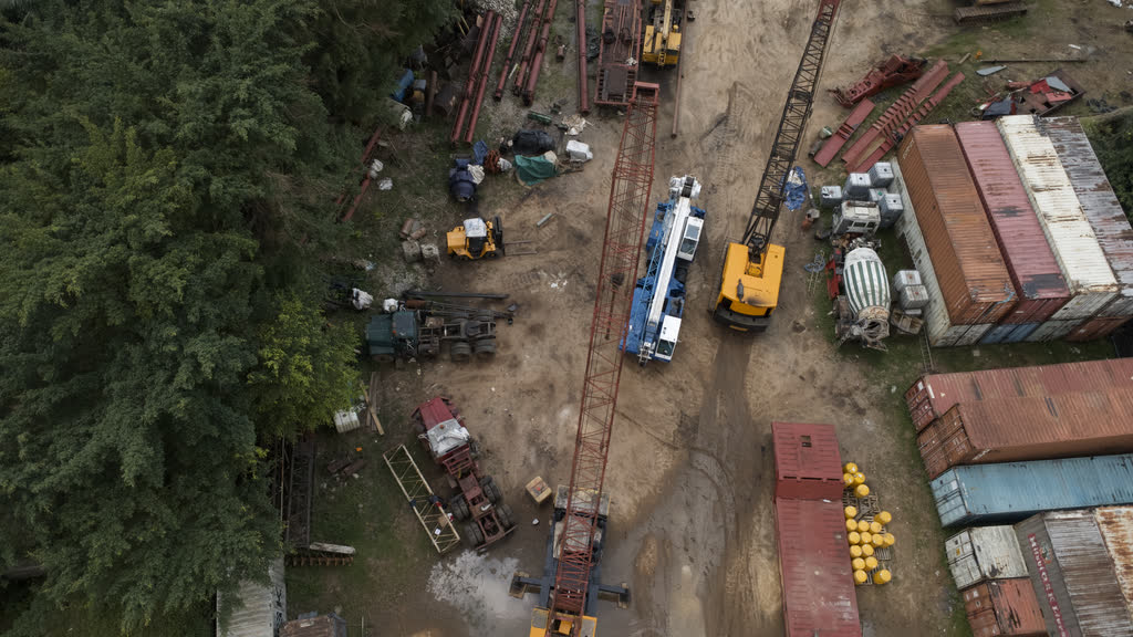 Overhead drone view of the equipment yard: crawler cranes, a piling rig, stacked steel sections, shipping containers and a line of trees