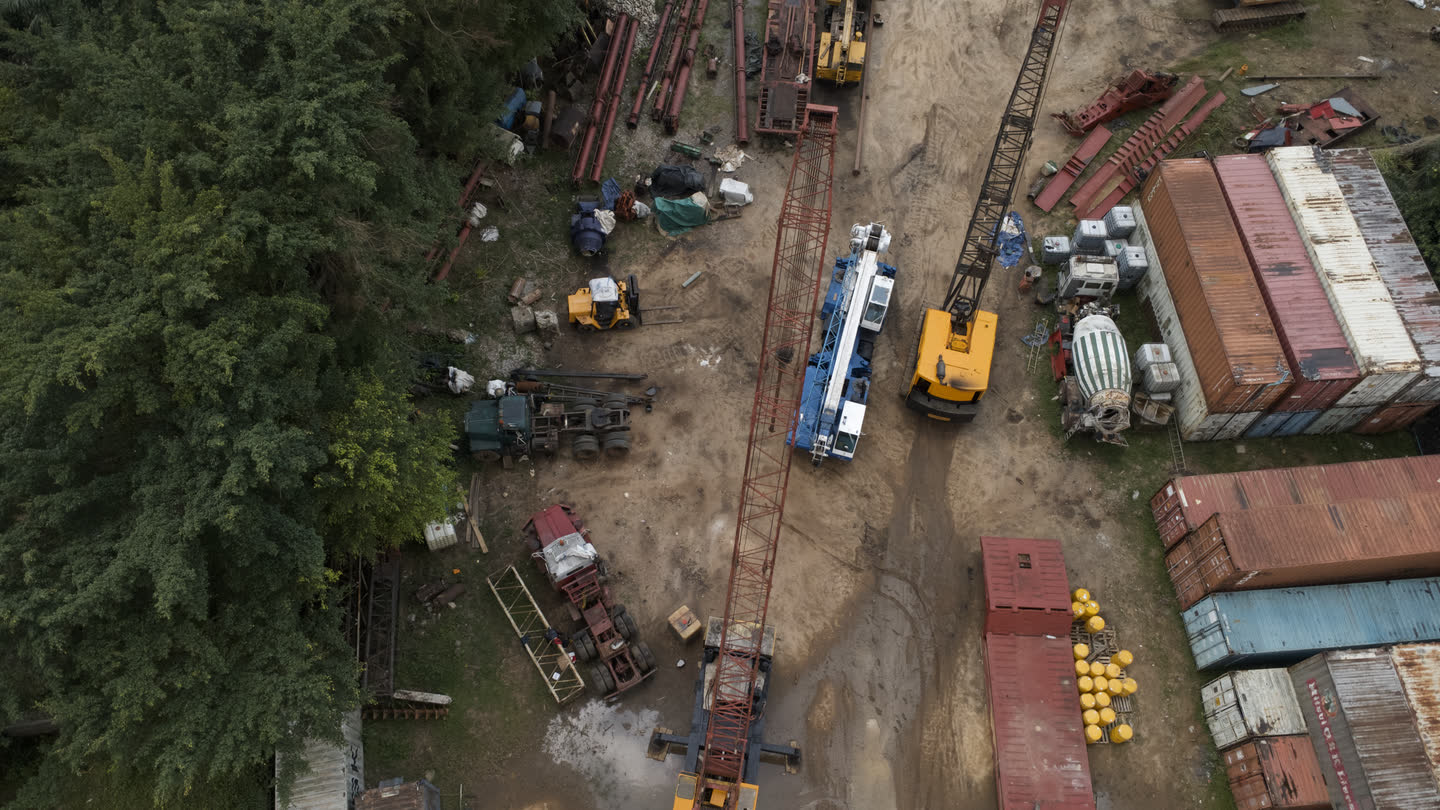 Overhead drone view of the equipment yard: crawler cranes, a piling rig, stacked steel sections, shipping containers and a line of trees