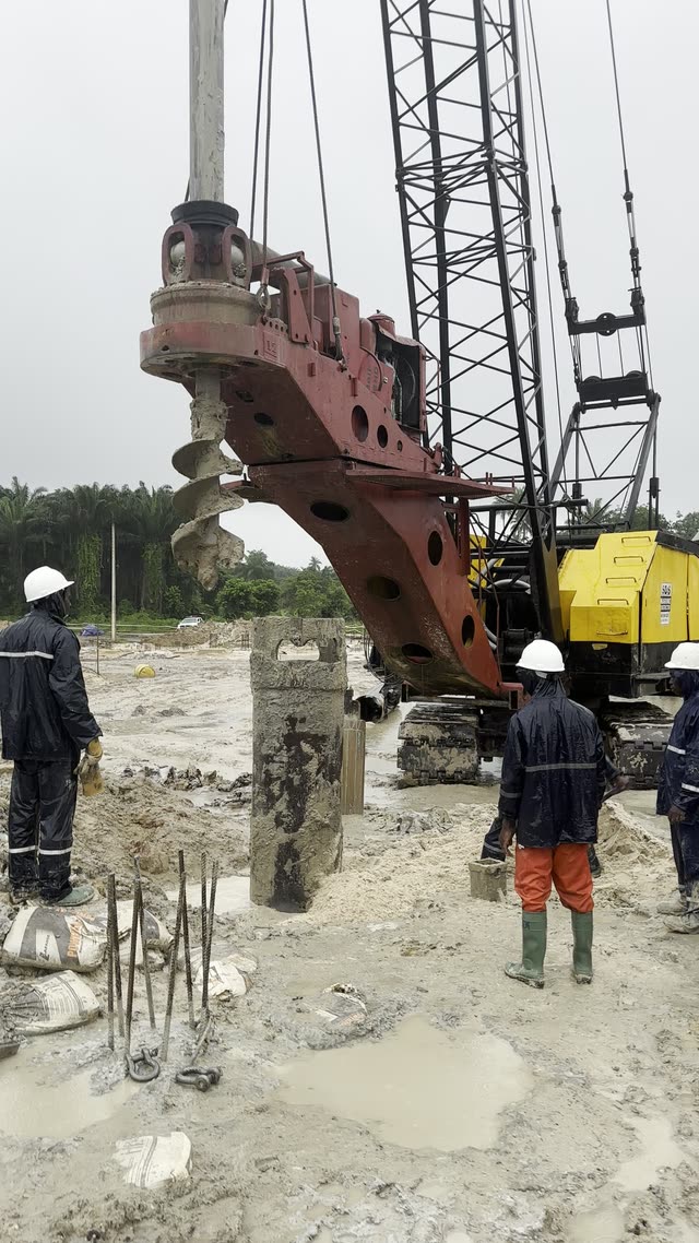 A red rotary piling rig turning an auger into saturated ground while two crew in coveralls watch from the pile position