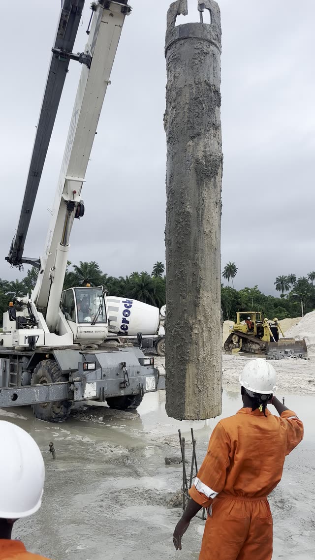 A mobile crane lifting a concrete-filled steel casing clear of the ground while a crew member in orange coveralls watches from the pile position