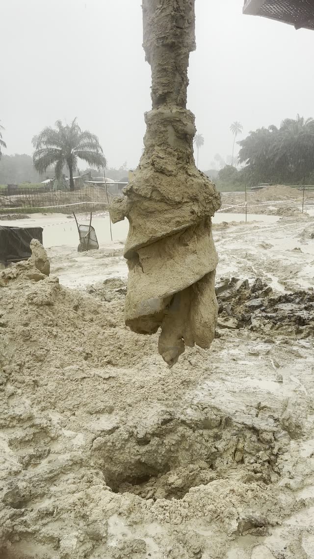 Close footage of a continuous-flight auger being withdrawn from a bore in heavy rain, wet spoil turning up the flights