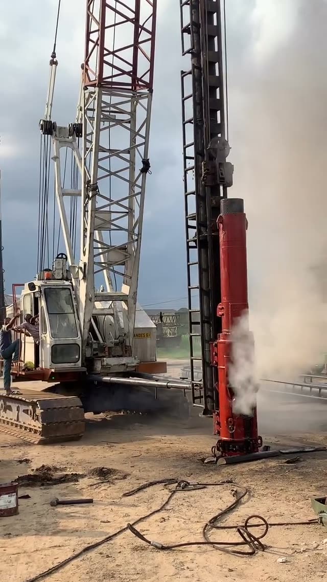A red diesel impact hammer firing in the leads of a crawler crane, each blow throwing a plume of exhaust across the site