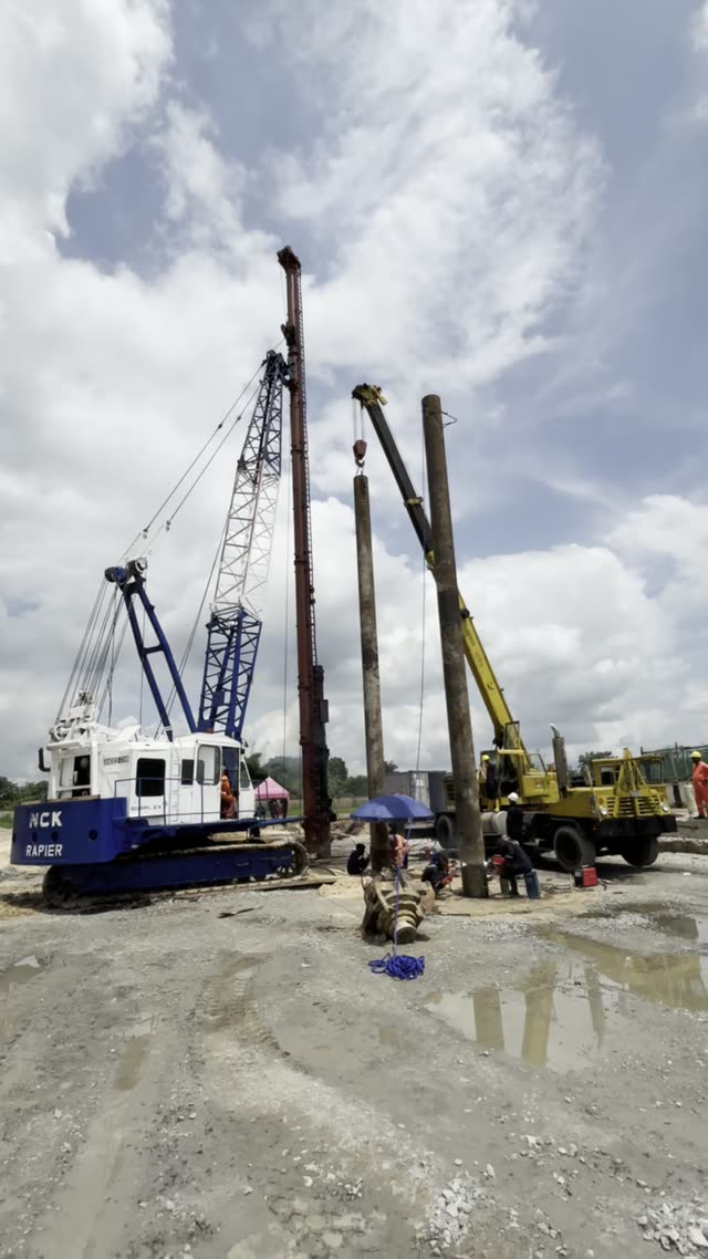 An NCK Rapier crawler crane standing with pitched steel piles against a bright sky of broken cloud, reflected in standing water