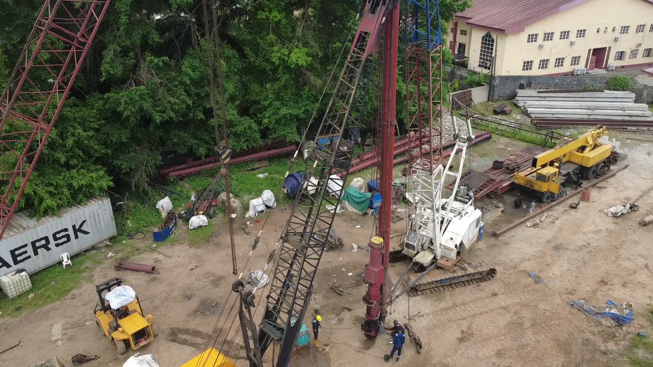 Aerial footage orbiting a red lattice-boom crane rigged with piling leads, with a service crane, stacked steel and containers around it