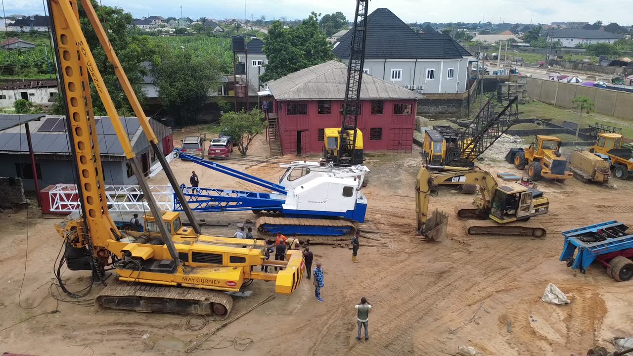 Aerial footage descending over the equipment yard: a large yellow rotary piling rig, a blue and white crawler crane, an excavator and crew at work